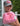 A woman wearing a gray 'Lucky' baseball cap holds up a pink chunky knit purse decorated with pastel pink, peach, and  sequins, set against a background of a house exterior and green shrubs.