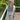 A woman wearing a white sleeveless top and light blue jeans holds up a large gray macrame fringe bag in front of her face, standing on a wooden deck with green trees in the background.