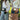 Two women standing outdoors near a tree, each wearing a crossbody bag with a heart-shaped cutout. The top bag is brown with a white heart, and the bottom bag is black with a white heart.