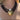 A close-up of a model wearing a bohemian-style multi-strand necklace. The necklace features three dark brown beaded strands, a large pearl bead, and a large, textured, faceted gold heart-shaped pendant.