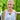 A woman with blonde hair in a bun stands on a wooden deck wearing a sleeveless white top with 3D rose appliqués and a colorful butterfly necklace, with green hedges in the background.