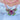 A close-up of a layered necklace worn on a person's neck, featuring a pink beaded top layer and a central butterfly pendant with pink and green stones. The necklace is styled over a white rose against a light blue background.