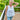 A woman wearing a white sleeveless top with 3D floral appliques and light blue jeans, standing on a wooden deck with green trees in the background.